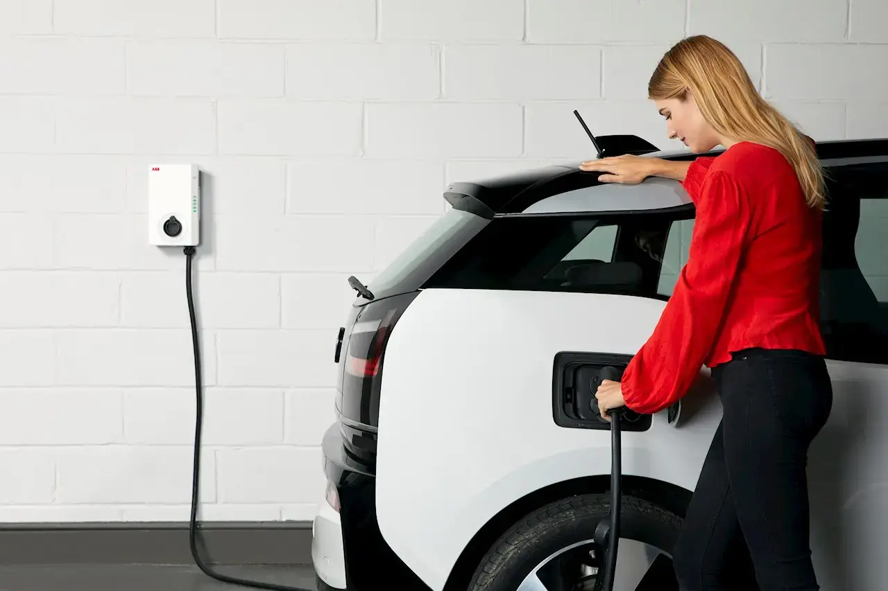 Person in a red long-sleeve top plugging a white electric vehicle with a black roof into a wall-mounted charging station attached to a white brick wall.