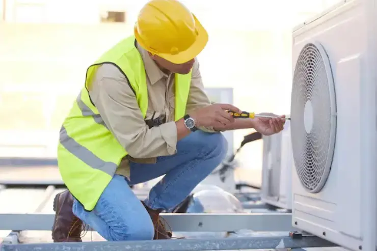 A worker in a yellow hard hat, safety vest, and work boots kneels on a rooftop, using a screwdriver to repair an outdoor air conditioning unit with a visible fan grille.