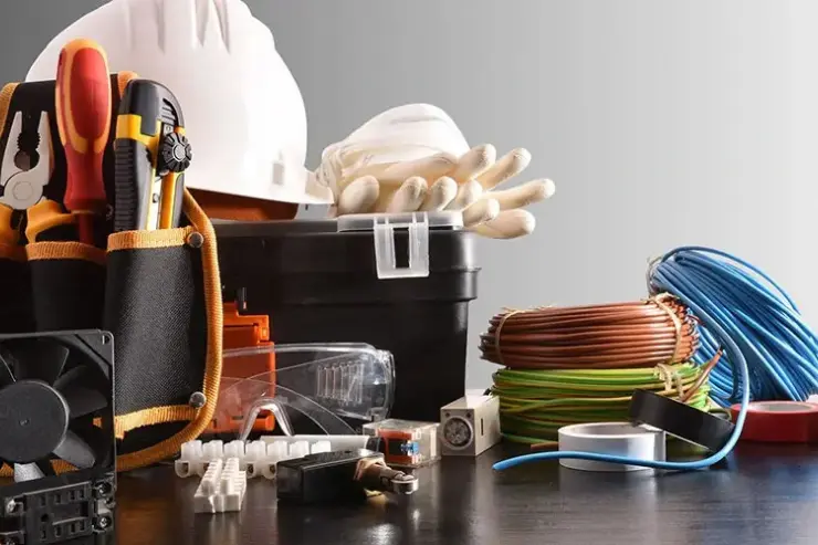 An assortment of electrical tools and equipment displayed on a dark surface, including a tool pouch with pliers and screwdrivers, a white safety helmet, gloves, goggles, electrical wires, tape rolls, and connectors.