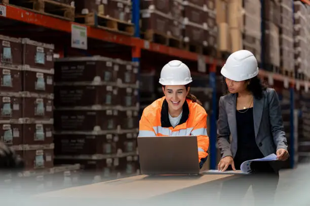Two individuals wearing white safety helmets stand at a workstation in a warehouse; one in an orange high-visibility jacket works on a laptop, while the other in a dark blazer holds rolled-up blueprints, with shelves of large boxes in the background.