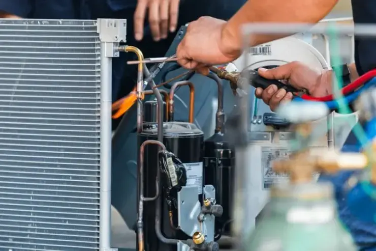 Technician using a torch to solder copper tubing connections on an HVAC system near a compressor unit, with visible pipes, refrigerant lines, and servicing tools.