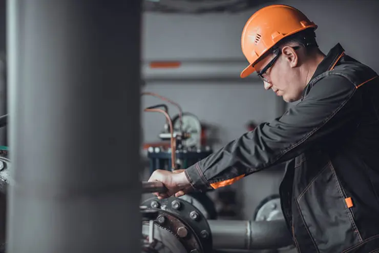 Worker wearing a black dress and orange colour safety helmet and protective glasses adjusting a valve on industrial piping inside a factory setting, with metal pipes and machinery in the background.