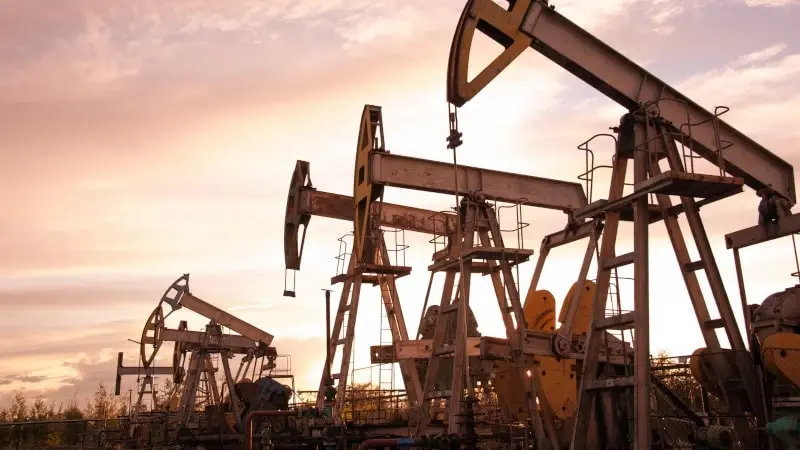 Multiple oil pumpjacks operating in a field during sunset, silhouetted against an orange and pink sky.