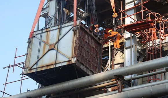 wo workers in safety gear guiding a heavy rusted rectangular structure suspended by steel cables in an industrial plant.