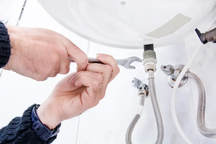 Hands using a wrench to adjust a pipe connection beneath a water heater, with braided hoses and metal fittings visible in a clean indoor setting.<br />
