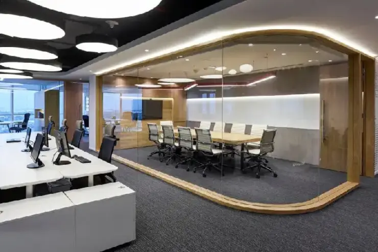 A sleek office space divided into two areas. On the left, an open workspace features desks with computer monitors and ergonomic chairs. On the right, a glass‑walled conference room contains a long wooden table surrounded by chairs, a large wall‑mounted display screen, and a whiteboard surface. Circular ceiling lights and wood accents create a professional yet modern atmosphere.