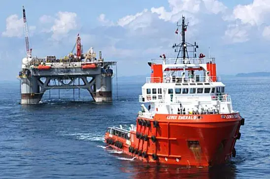 A large red offshore supply vessel named “Lewek Emerald” sails near a semi-submersible oil drilling platform equipped with cranes and machinery, under a partly cloudy sky.brief it
