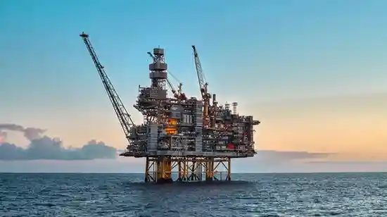 Offshore oil drilling platform in the ocean, supported by steel legs and equipped with cranes, pipes, and industrial machinery, illuminated against a sunset sky.
