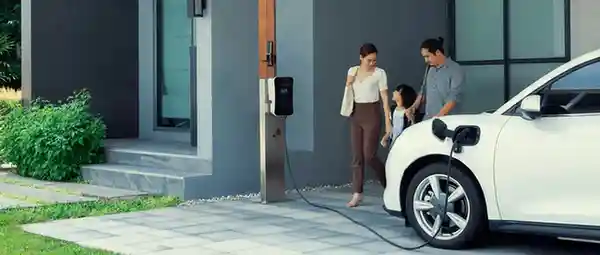 A white electric car is parked in a modern driveway, connected to a home charging station. Three people stand nearby, interacting in front of a contemporary house with large windows.