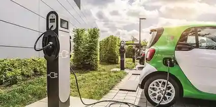 A small green and white electric car is charging at an outdoor station. Another EV and multiple chargers are visible in the background, with a modern building and greenery under a partly cloudy sky.