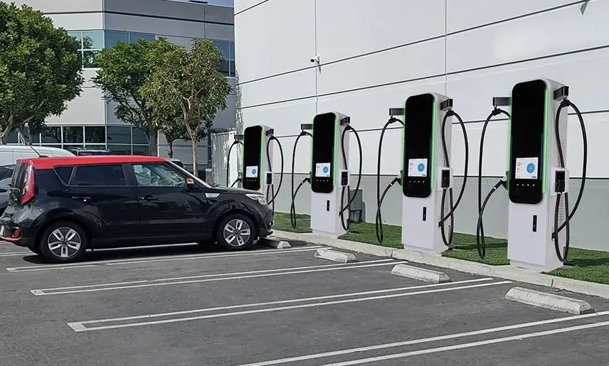 Five tall electric vehicle charging stations line a building wall in a parking lot. A black and red compact EV is plugged in, while large windows and trees frame the background.