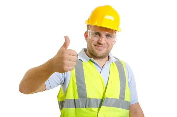 A person wearing a yellow hard hat, clear safety glasses, and a high-visibility vest with reflective stripes stands against a plain white background, smiling and giving a thumbs-up gesture to convey workplace safety and confidence.