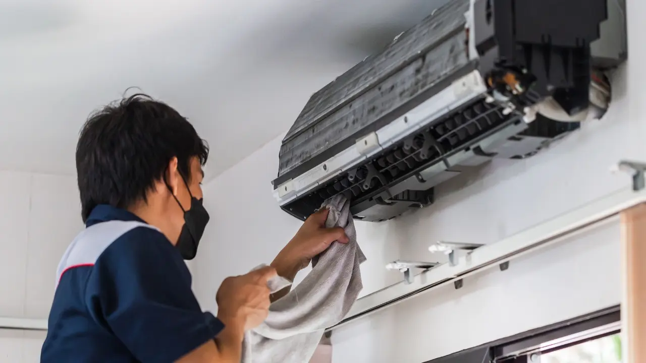 A person in a dark shirt and black face mask cleans the interior of a wall-mounted air conditioner, wiping exposed evaporator coils and filters with a cloth.
