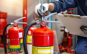 A person wearing gloves inspects red fire extinguishers during a safety check, holding a clipboard while examining the pressure gauge and safety pin.