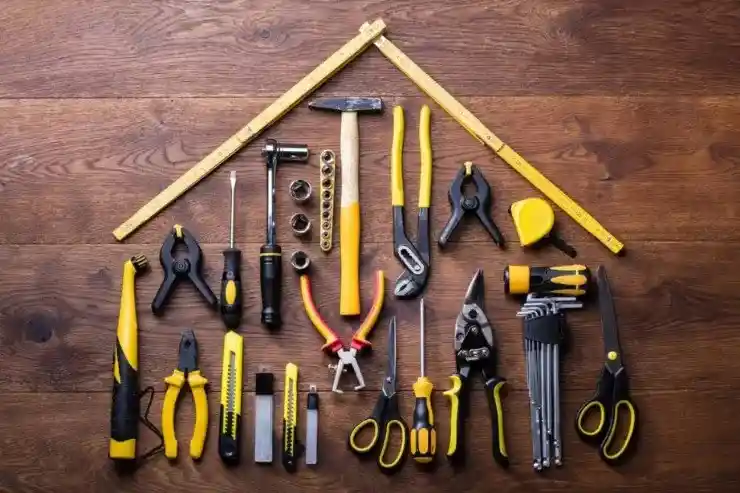 "Assorted black-and-yellow hand tools arranged on a wooden surface in the shape of a house, with a folding ruler forming the roof and items like a hammer, pliers, screwdrivers, wrenches, tape measure, and utility knives outlining the structure, symbolizing home improvement.
