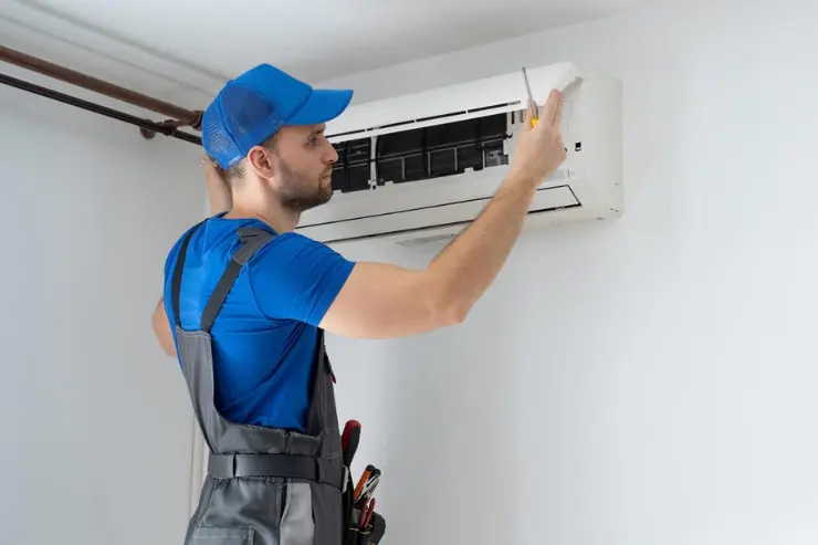 Person in blue cap, blue shirt, and gray overalls using a screwdriver to repair a wall-mounted air conditioning unit with its front cover open, exposing filters and coils. The indoor setting includes a white wall and curtain rod, illustrating HVAC maintenance and technical servicing.
