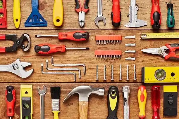 Neatly arranged collection of hand tools on a wooden surface, including hammer, wrenches, screwdrivers, pliers, Allen keys, tape measure, spirit level, paintbrush, utility knife, caliper, tin snips, drill bits, and screws, organized to display a complete toolkit for carpentry, mechanical, and household repair tasks