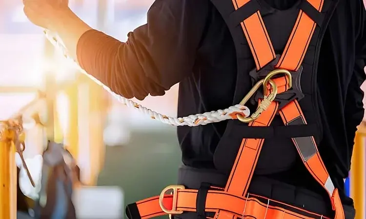 Worker wearing bright orange safety harness with black padding, clipped to a white braided safety rope using a metal carabiner. Industrial background with yellow railings and sunlight.